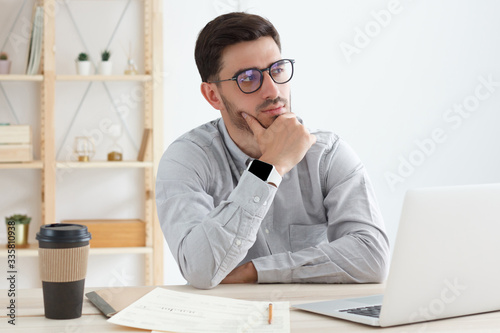 Young man in office wearing glasses, gray shirt, smart watch, tinking, scratching his chin, trying to find good idea for business optimization and improvement photo