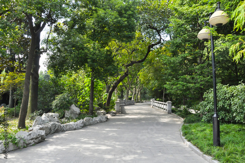 A concrete path through a park in the French concession in Shanghai, China. The path is surrounded by trees and lanterns and leads to a small white bridge.