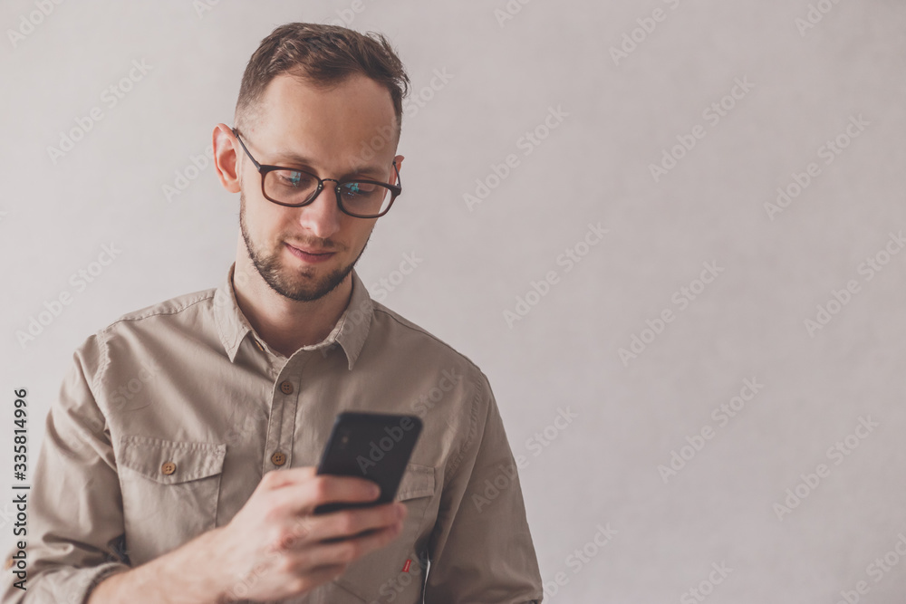 Joyful caucasian man dressed in beige shirt and wearing eyeglasses using cellphone on gray background. Looking at phone. Focus on face. Joyful caucasian man dressed in beige shirt and wearing eyeglasses using cellphone on gray background. Looking at phone. Focus on face.