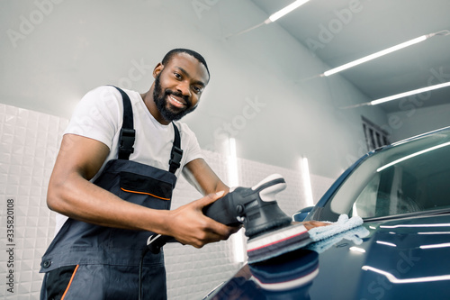 Smiling handsome young black skinned man, professional worker of car detailing service, polishing blue car with polishing machine and microfiber cloth. Car detailing, polishing, finishing concept