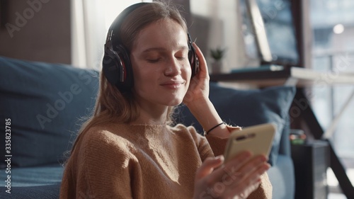 Shoot of cheerful european girl sittin on the floor near the sofa with headphones and listening music and looking into the phone.
