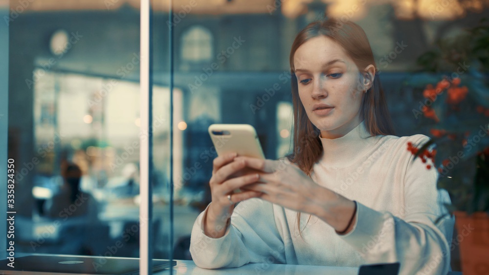 View through the window of attractive serious young woman using cellphone or smartphone at coffee shop, texing message, watching video or bloggs.