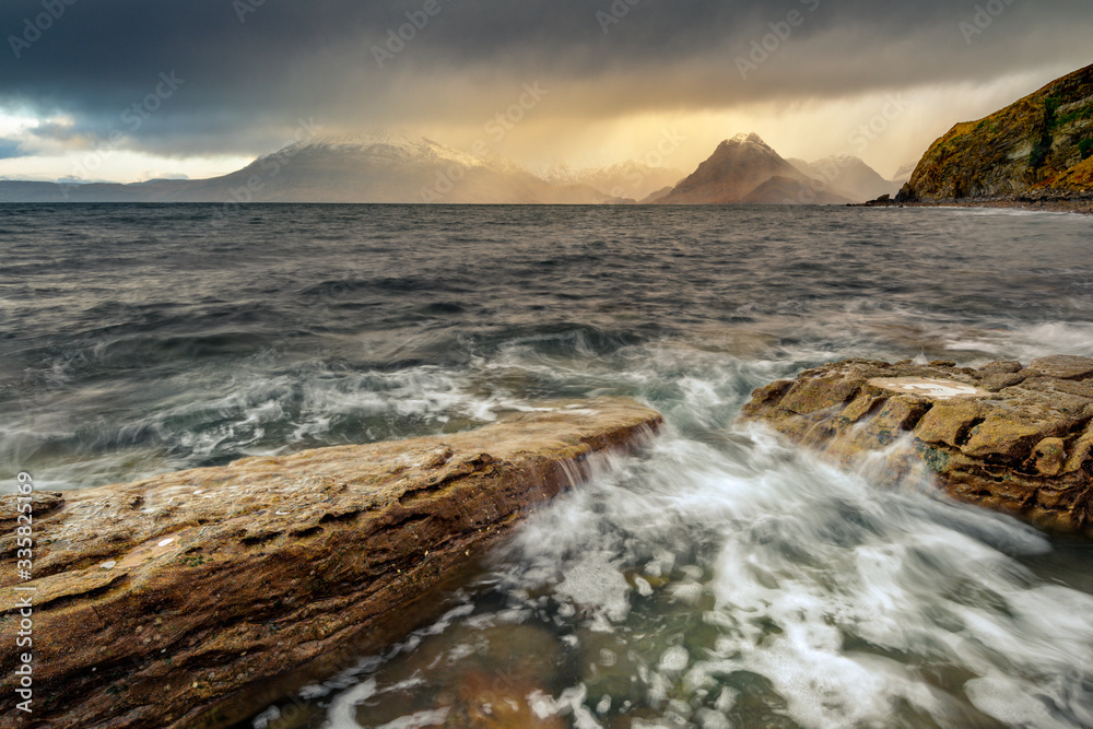 Dark And Moody Clouds With Rain Showers Passing Over Scottish Highland Mountains With Waves Crashing Over Rocks On Shoreline. Elgol, Isle Of Skye, UK.
