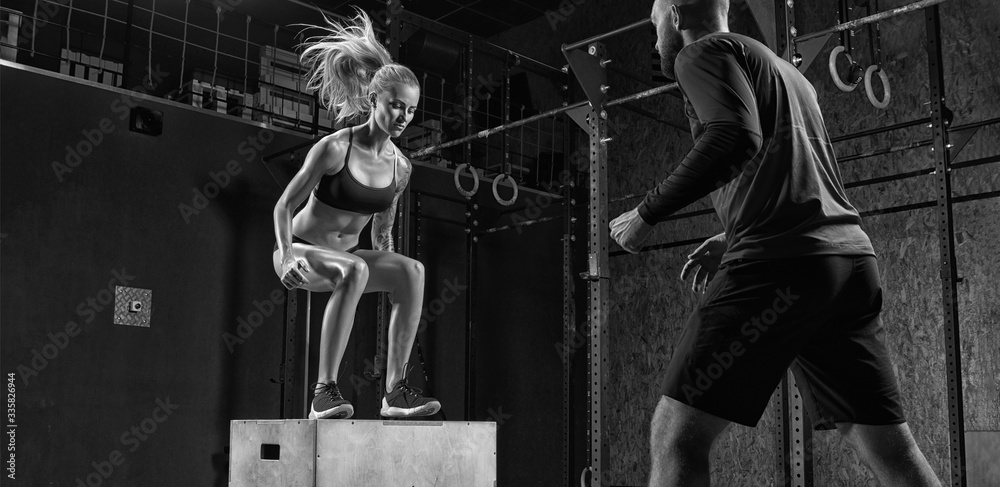 Fototapeta premium Fitness woman doing a box jump at the gym with the help of a personal trainer in a modern gym Trainer helping woman on her work out routines. A couple of athletes. Black and white photo.