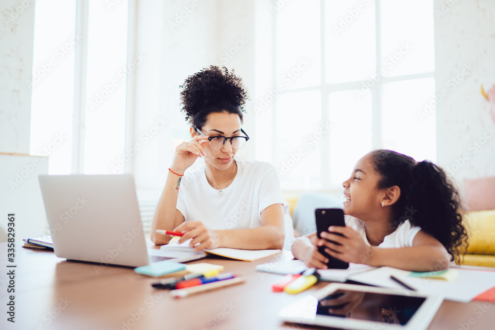 Mother using laptop and laughing daughter playing on smartphone while looking at each other Mother using laptop and laughing daughter playing on smartphone while looking at each other