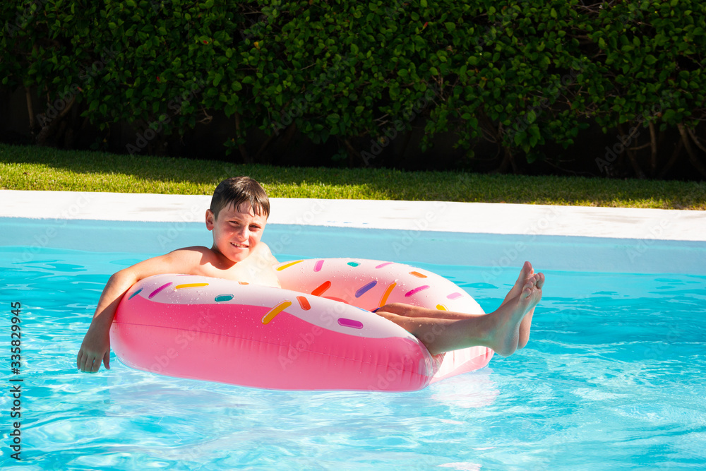 Smiling boy floating on rubber ring at apartment complex swimming pool on sunny day. Happy child enjoying vacation on pink float outdoors. Summer holidays, resort attraction concepts