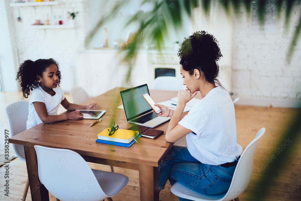 Fototapeta premium Black mother using smartphone and daughter playing on tablet