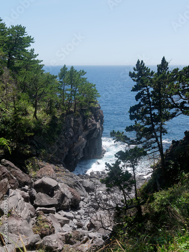 View of volcanic rocky inlet beach with boulders and pebbles in the Jogasaki coast in Izu, Japan. Beautiful ocean is seen between two cliffs with pine trees. Waves crushing against rocky shore.
