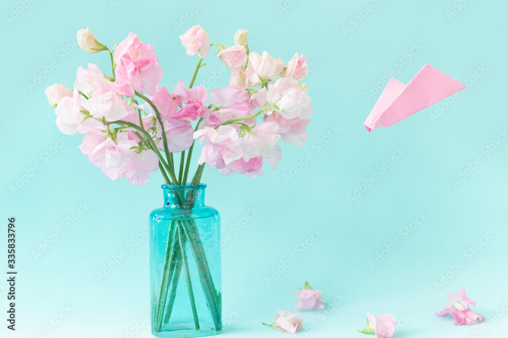 Obraz premium Bouquet of pink sweet pea Lathyrus in beautiful transparent vase on blue background.Paper airplane with message. Spring concept. International Women's Day, March 8 and Valentine's Day,14 of February