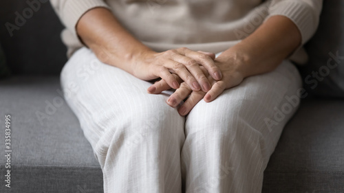 Close up elderly senior woman sitting on sofa, holding folded hands on lap, feeling lonely at home. Middle aged mature lady suffering from depression, recollecting memories alone in living room.