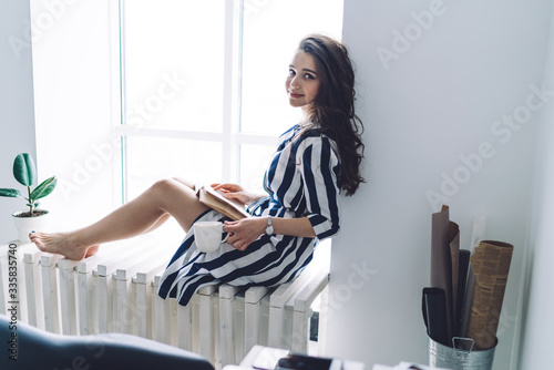 Pleasant pretty woman reading book on window sill at home