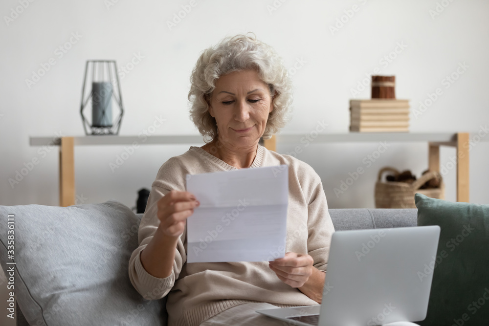 Pleasant smiling elderly mature woman sitting on sofa, holding laptop on lap, reading banking loan paper notification. Happy older senior grandmother looking through mail letter alone at home. Pleasant smiling elderly mature woman sitting on sofa, holding laptop on lap, reading banking loan paper notification. Happy older senior grandmother looking through mail letter alone at home.