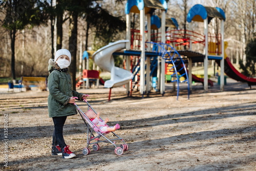 Sad little girl in a protective mask on the background of the closed playground outdoor. A child in a medical mask walks in the park. Quarantine measures against COVID-19. Coronavirus pandemic.