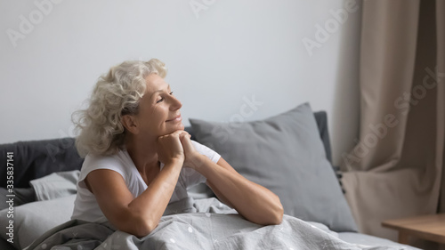 Foto Peaceful calm positive middle aged senior retired woman sitting in bed after wakeup in weekend morning, enjoying good mood after good night rest relaxation, welcoming new day alone in bedroom