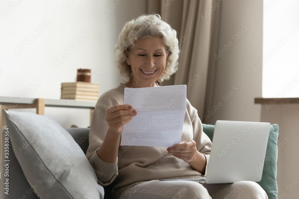 Happy middle aged elderly woman sitting on couch, holding computer on knees lap, reading paper document or mail letter from friend. Smiling mature old grandmother excited by good news notification. Happy middle aged elderly woman sitting on couch, holding computer on knees lap, reading paper document or mail letter from friend. Smiling mature old grandmother excited by good news notification.