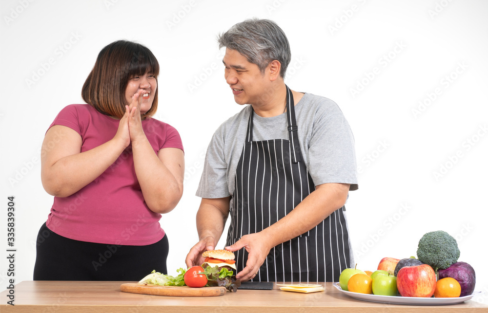 Happy overweight couple cooking a burger in the kitchen room. A vegetable on the table. Concept of happy family and binge eating Happy overweight couple cooking a burger in the kitchen room. A vegetable on the table. Concept of happy family and binge eating
