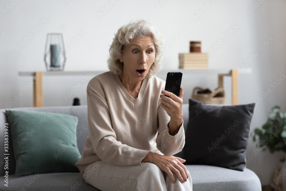 Shocked middle aged grey haired woman resting on sofa, looking at smartphone screen, reading message, surprised by unbelievable news. Excited mature granny received online lottery win notification. Shocked middle aged grey haired woman resting on sofa, looking at smartphone screen, reading message, surprised by unbelievable news. Excited mature granny received online lottery win notification.