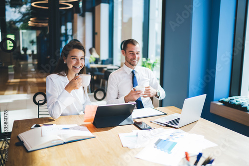 Cheerful male and female entrepreneurs enjoying live communication for business brainstorming in team sitting at desk table in office interior and smiling, happy colleagues have coffee break