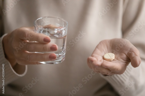 Close up senior mature woman holding glass of fresh pure distilled water and round pill. Unhealthy elderly grandmother taking painkiller, relieving headache. Healthy granny taking healthcare vitamins.
