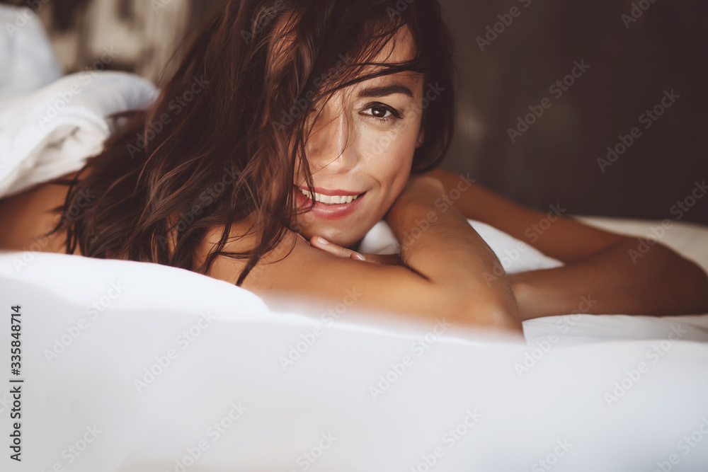 Obraz premium Close-up portrait Beautiful woman in the morning in a white bed after waking up, smiles and covers her face with hair. Soft selective focus. Toned photo.