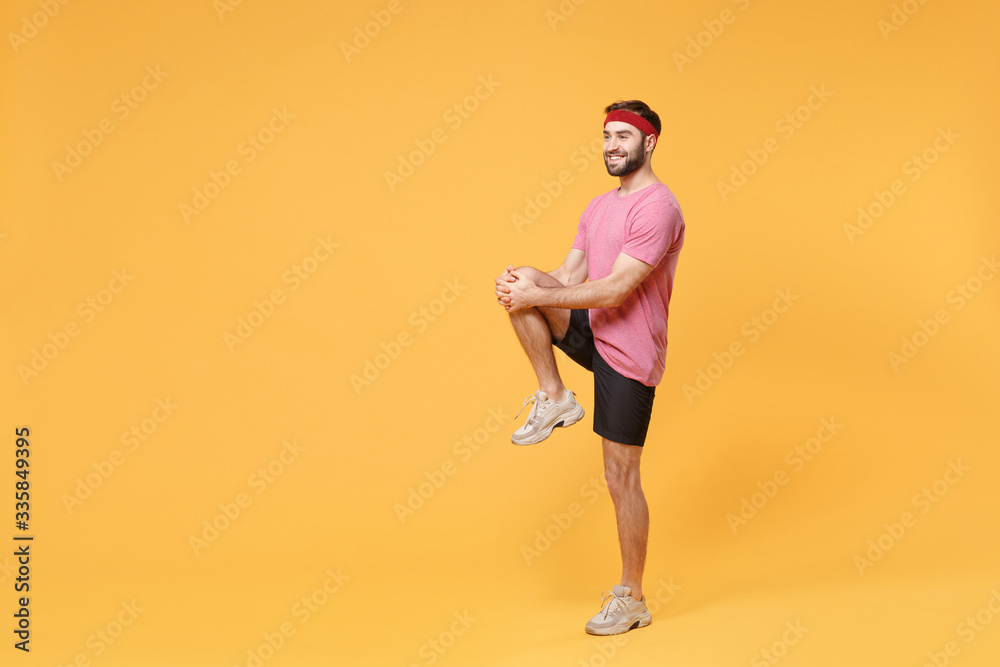 Smiling young bearded fitness sporty guy 20s sportsman in headband t-shirt in home gym isolated on yellow background. Workout sport motivation concept. Doing warm up, stretching exercising for legs.