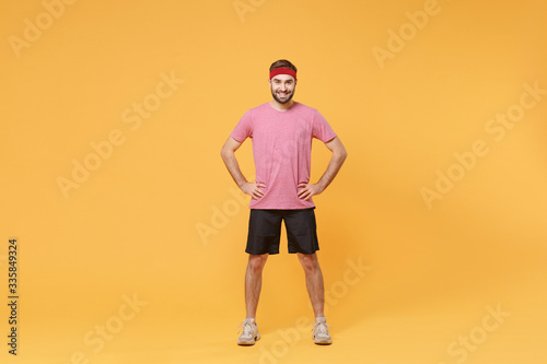 Smiling young bearded fitness sporty guy sportsman in headband t-shirt spend weekend in home gym isolated on yellow background. Workout sport motivation concept. Standing with arms akimbo on waist.