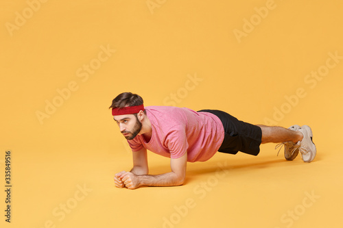 Young bearded fitness sporty guy 20s sportsman in headband t-shirt spend weekend in home gym isolated on yellow background in studio. Workout sport motivation lifestyle concept. Doing plank on floor.