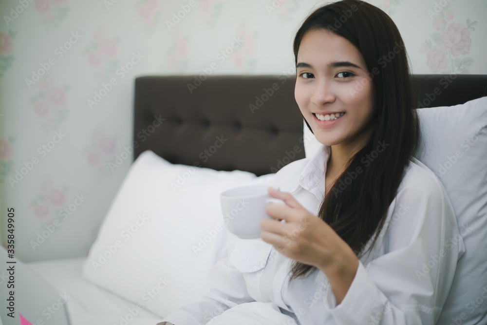 Asian woman is drinking a cup of coffee and using a notebook to work on the bed.She smiles and enjoys working at home. Asian woman is drinking a cup of coffee and using a notebook to work on the bed.She smiles and enjoys working at home.