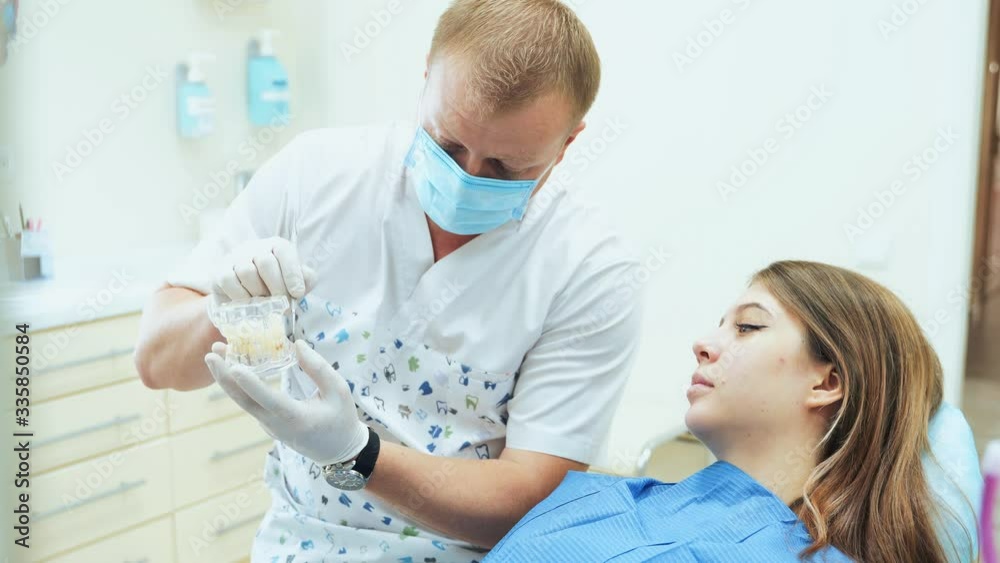 A doctor in a mask and rubber gloves demonstrates a mock jaw to a patient. A girl at the dentist's appointment in the clinic. A doctor in a mask and rubber gloves demonstrates a mock jaw to a patient. A girl at the dentist's appointment in the clinic.