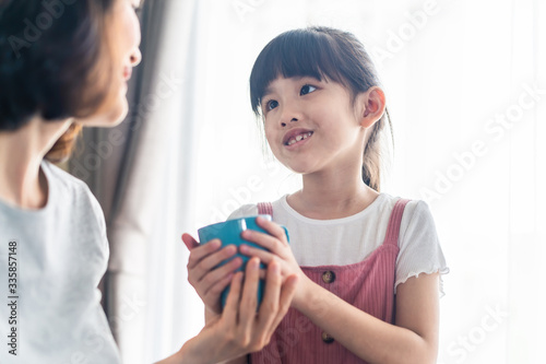 Asian young office mother work from home during coranavirus outbreak. Cute daughter give a cup of coffee to mom and kiss for a break and rest. Love care in family. Covid-19, Social distance concept