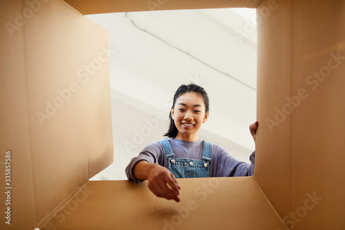 Low angle view at smiling Asian woman reaching into cardboard box while packing or unpacking for new home, copy space photo