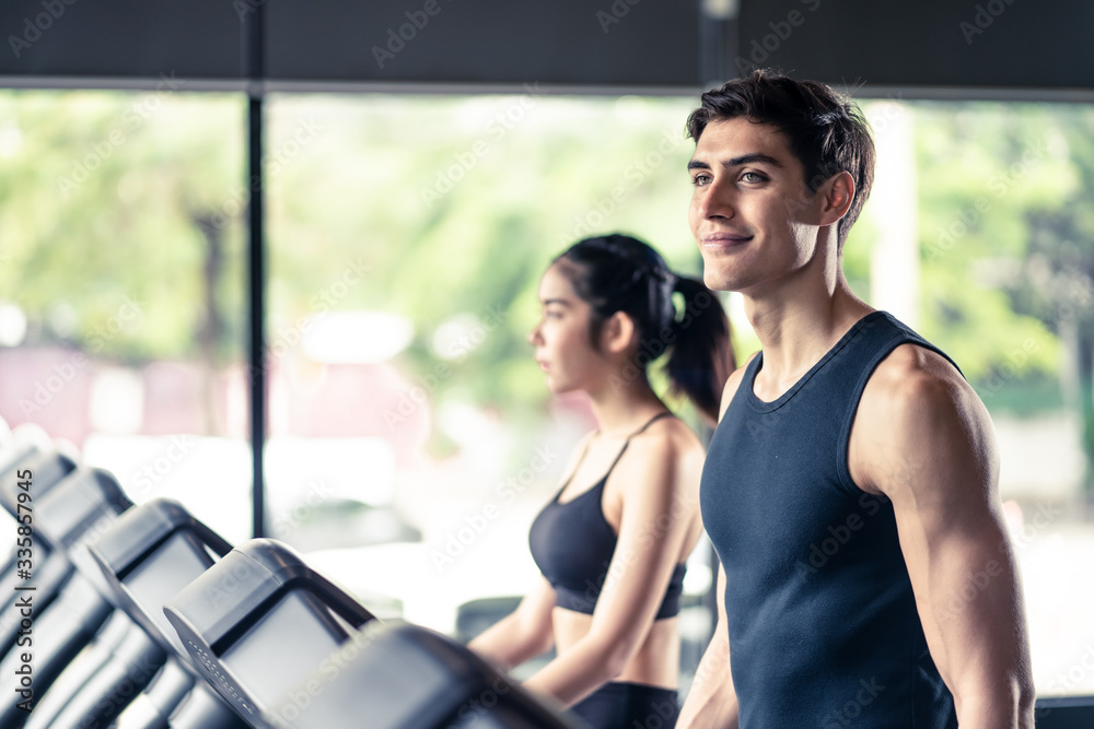 Young fit beautiful Asian woman and handsome Caucasian man walking on treadmill or running machine in modern fitness gym. Seen from side view while they focusing on running. Workout in Gym and fitness