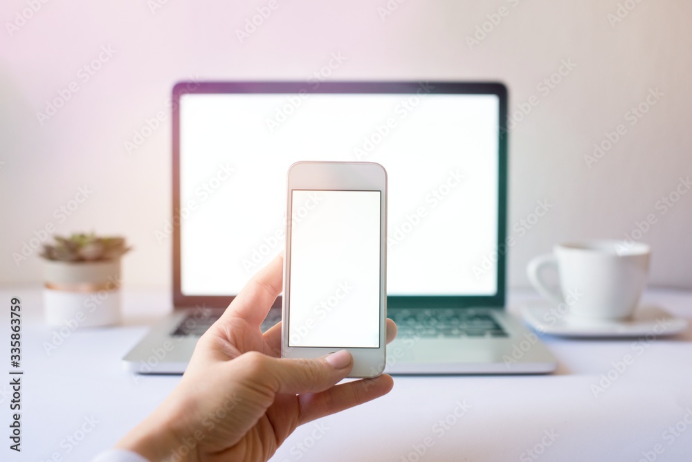 Close-up of smartphone with blank screen in hands of young woman sitting at white table and touching screen. On table laptop with blank screen,cup of coffee. White style. Minimal. Mock up,space for ad Close-up of smartphone with blank screen in hands of young woman sitting at white table and touching screen. On table laptop with blank screen,cup of coffee. White style. Minimal. Mock up,space for ad