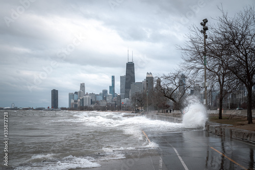 Photo Damaging waves crash against the shoreline and erode the lakefront bike path or trail with Chicago skyline in background on a cloudy and windy day