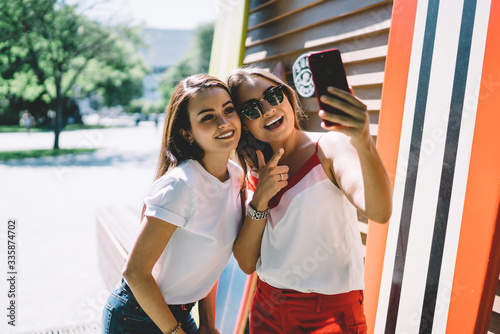 Happy trendy millennial women taking selfie with smartphone while resting together in park