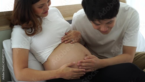 young asian  pregnant woman sitting on bed and husband touch the bally expecting baby with love during home quarantine from covid-19 virus pandemic photo