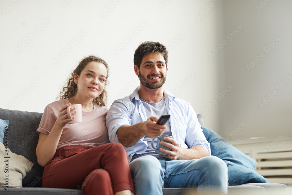 Portrait of modern couple watching TV and holding mugs while sitting on sofa at home in cozy apartment enjoying lazy time, copy space Portrait of modern couple watching TV and holding mugs while sitting on sofa at home in cozy apartment enjoying lazy time, copy space