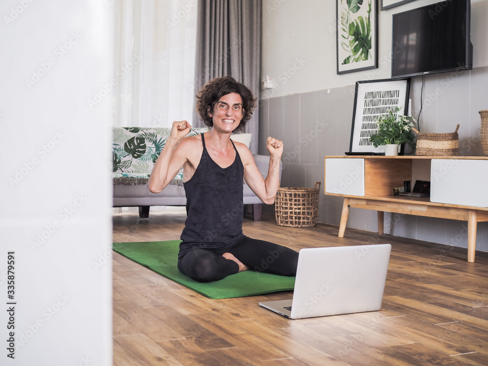 happy excited and smiling women in winner pose sitting crossed legged on the floor on a yoga mat in her living room at home looking at laptop having a video conference 