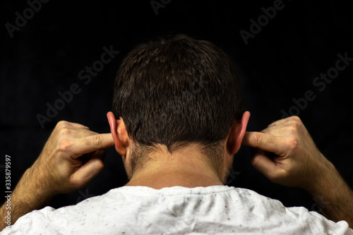 A man in a white T-shirt covers his ears with his fingers. Close up on black background. Back view.