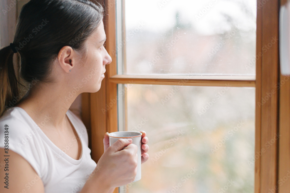 Interior shot of young female holding a cup of tea looking at the outside world through the window gazing into the distance. Interior shot of young female holding a cup of tea looking at the outside world through the window gazing into the distance.