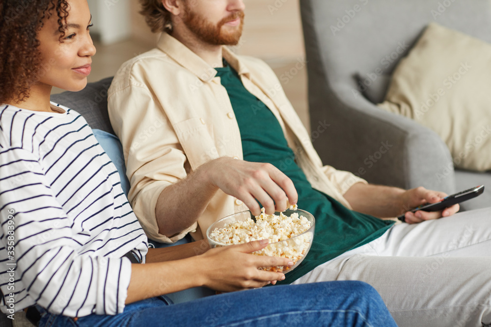 Cropped portrait of modern mixed-race couple watching TV at home while relaxing on cozy sofa, focus on hands holding bowl of popcorn, copy space Cropped portrait of modern mixed-race couple watching TV at home while relaxing on cozy sofa, focus on hands holding bowl of popcorn, copy space