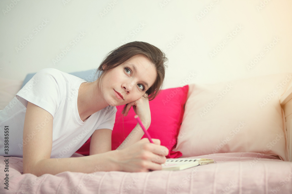 Young woman writing something in the notebook on the bed. Peaceful atmosphere. Woman stay home during a quarantine Young woman writing something in the notebook on the bed. Peaceful atmosphere. Woman stay home during a quarantine