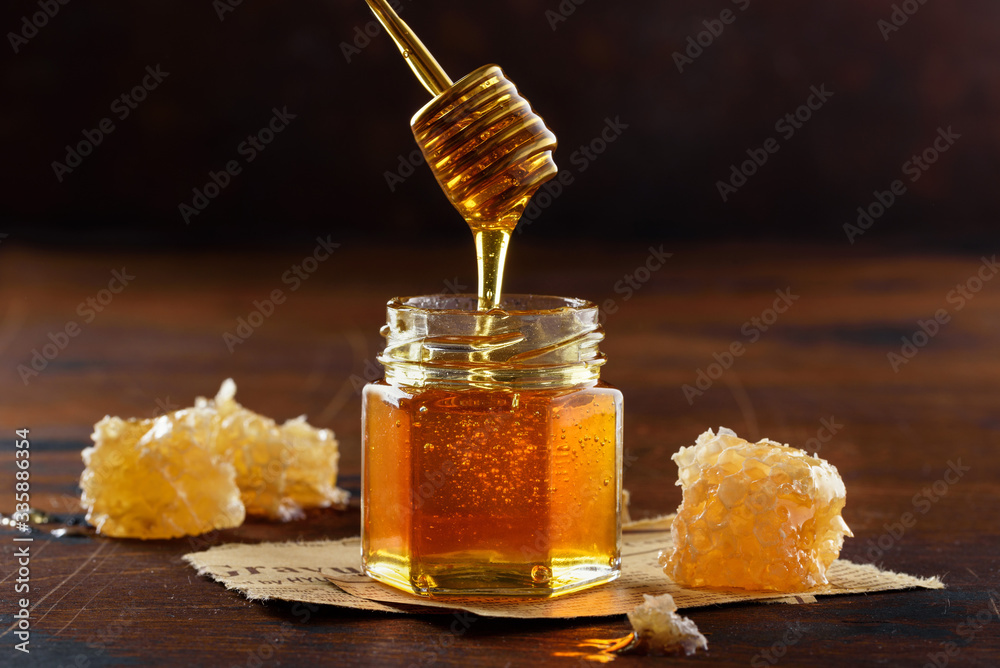Glass jar and honey dripping from dipper on dark wooden background. Honeycomb. Selective focus Glass jar and honey dripping from dipper on dark wooden background. Honeycomb. Selective focus
