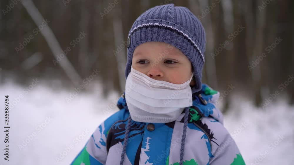 A child (boy) in winter overalls and a hat stands in the winter forest with a medical mask on his face, looks at the camera and removes the mask from his face, the end of the epidemic and self-isolati A child (boy) in winter overalls and a hat stands in the winter forest with a medical mask on his face, looks at the camera and removes the mask from his face, the end of the epidemic and self-isolati