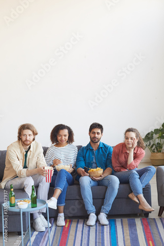 Vertical full length portrait of multi-ethnic group of friends watching TV together while sitting on comfortable sofa at home and enjoying snacks, copy space in top section