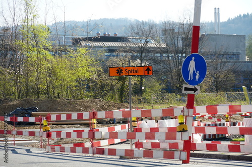 construction site of civil engineering with signs for deviation or detour for pedestrians and alternate route to hospital written in German, secured by barrier planks in red and white and lights.