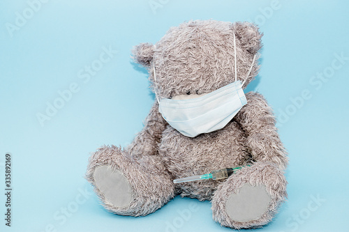 Toy grey bear sits on a blue background in a mask and holds a syringe