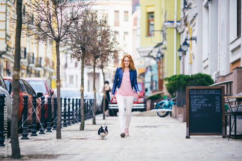 young redhaired Caucasian woman walking along European street with small Chihuahua breed dog of two colors on leash. Cloudy, warm autumn spring weather. Girl Dressed in leather jacket and pink shoes © Elizaveta