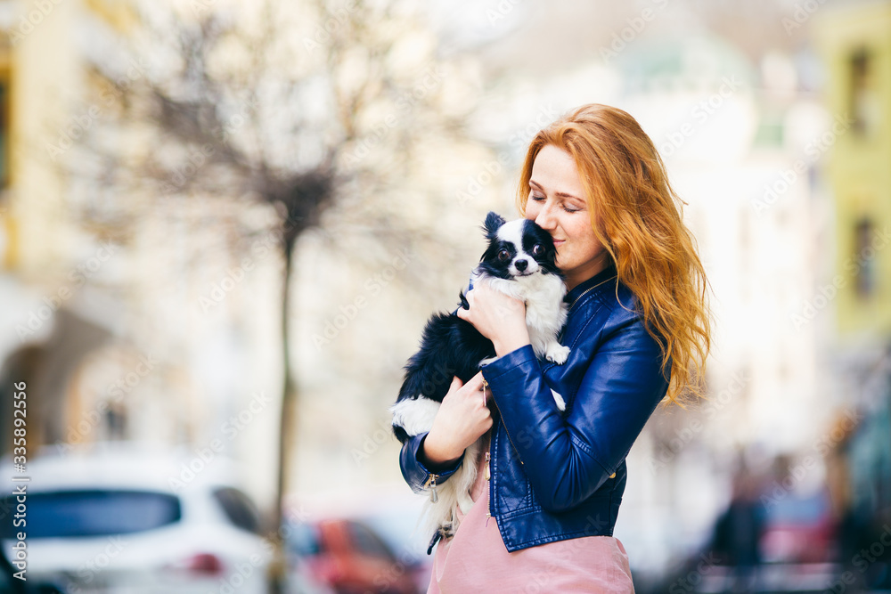 A young redhaired Caucasian woman with freckles holds and kisses, embracing black and white shaggy dog of Chihuahua breed. girl dressed in blue leather jacket, stands on street in spring in Europe