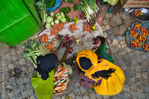 Fotografía Black women in colorful traditional African costumes crouching at a vegetable stand on the ground and buying vegetables